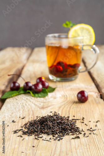 black tea in a glass cup with mint cherries and lemon on a wooden table next to fresh cherries and sprinkled tea leaves.