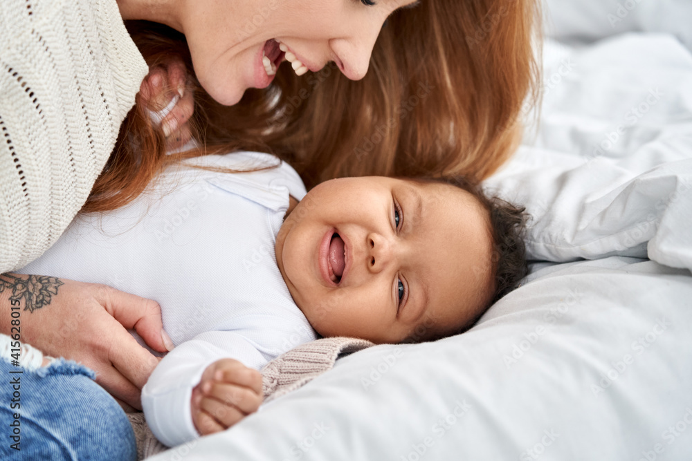 Happy young caucasian mother having fun with cute adorable little baby daughter on bed. Mixed race family diverse mum and funny african american infant child girl playing, laughing together at home. Happy young caucasian mother having fun with cute adorable little baby daughter on bed. Mixed race family diverse mum and funny african american infant child girl playing, laughing together at home.