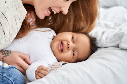 Happy young caucasian mother having fun with cute adorable little baby daughter on bed. Mixed race family diverse mum and funny african american infant child girl playing, laughing together at home.