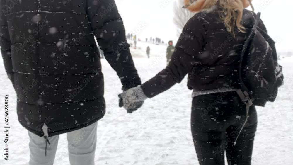 Beautiful young woman and man are holding hands in winter park under falling snow. Happy couple in love enjoys time together. Winter leisure. Beautiful young woman and man are holding hands in winter park under falling snow. Happy couple in love enjoys time together. Winter leisure.