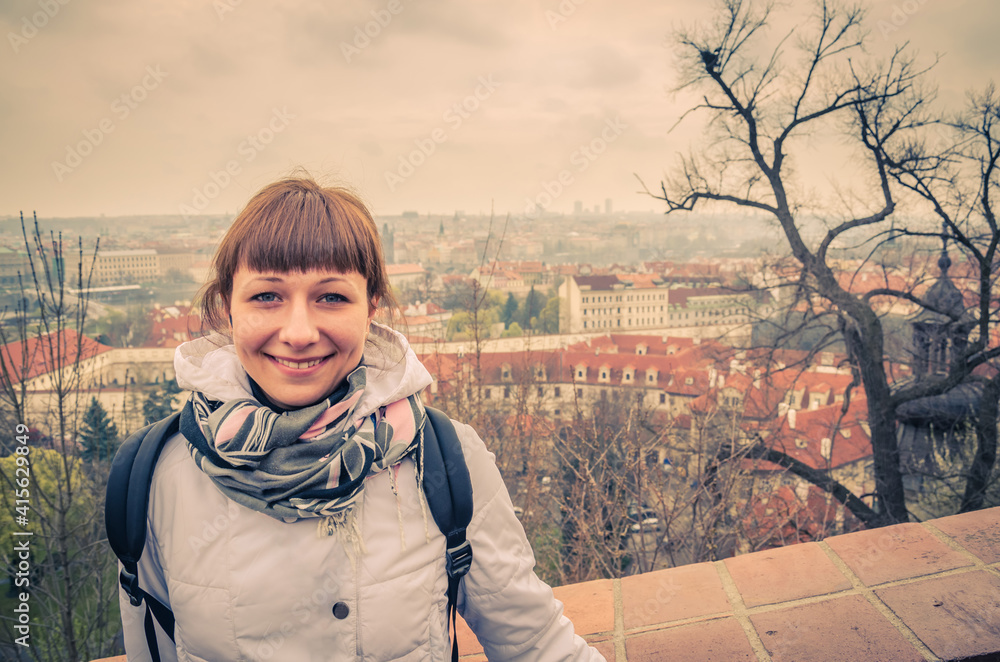 Close-up portrait of young caucasian girl traveler with white jacket looking at camera and smile, aerial view of Prague city historical centre background, woman on vacation in Czech Republic