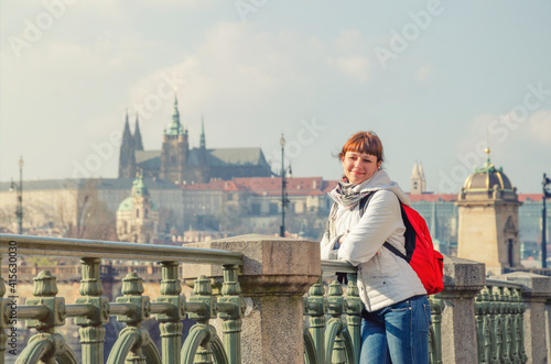 Photography Young girl tourist with white jacket and red backpack looking away and posing near fence of Vltava river embankment in Prague city historical centre, Prague Castle and St