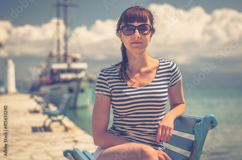 Young beautiful girl with striped t-shirt and sunglasses smile and sit on bench in pier, white ship on Toroneos gulf blue turquoise water in Halkidiki in sunny summer day background, close-up view photo