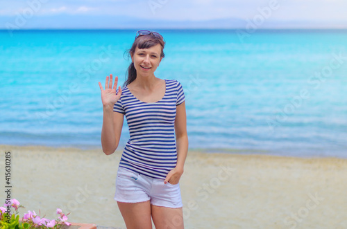 Young beautiful girl with striped t-shirt and white shorts posing and show hello on sandy beach, turquoise water of Toroneos gulf in Halkidiki Kassandra, sunny summer day, woman on vacation in Greece photo