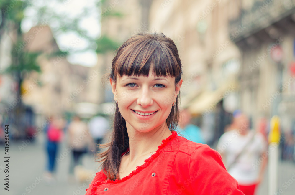 Close-up portrait of young beautiful caucasian girl with red jacket looking at camera and smile in pedestrian european city street with blurred background, woman with dark hair
