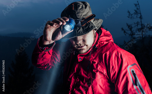 Photo of a male hiker in red jacket and panama putting on head mount flashlight on dark mountain forest background. photo