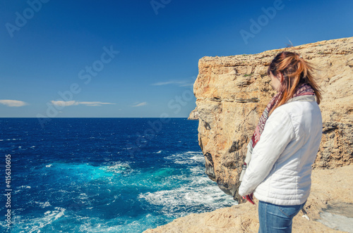 Young woman traveler with white jacket and headscarf blurred looking away distance on stone rocks of Dwejra Bay with collapsed Azure Window in Gozo island of Mediterranean sea, vacation on Malta
