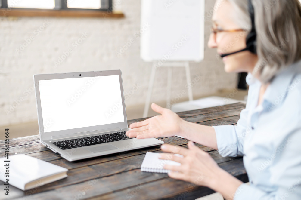 Senior gray-haired entrepreneur businesswoman teacher in headset sitting at the desk, meeting online with client, video calling via laptop from office, looking at the blank screen, presenting project Senior gray-haired entrepreneur businesswoman teacher in headset sitting at the desk, meeting online with client, video calling via laptop from office, looking at the blank screen, presenting project