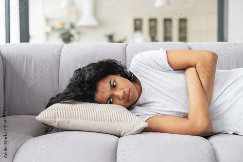 Angry African-American woman is lying on a white sofa arms crossed with sad, irritated and thoughtful face looking down, at home, after breaking up with boyfriend, cannot fly away due to quarantine