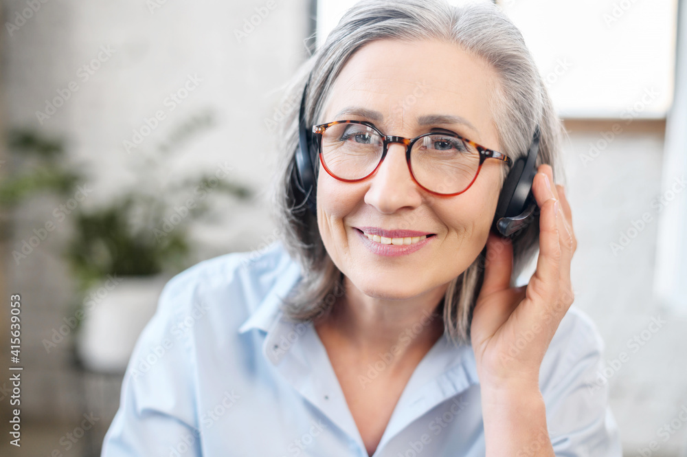 Headshot portrait of a charming smiling elegant senior lady, wearing headset, glasses looking at the camera. Portrait of a middle-aged gray-haired customer service representative sitting in the office Headshot portrait of a charming smiling elegant senior lady, wearing headset, glasses looking at the camera. Portrait of a middle-aged gray-haired customer service representative sitting in the office