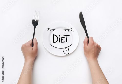 Female hands hold a fork and knife near a white plate on which the word Diet is written and a funny face is drawn.The concept of a balanced diet,ration, medical fasting.Top view,background, copy space © shchus