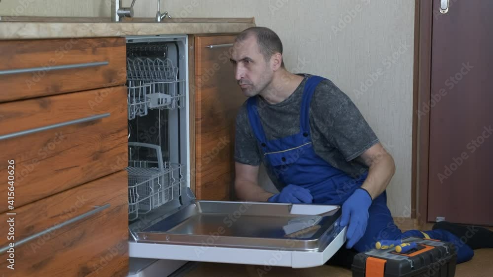 Dishwasher service. A man in overalls and tools repairs a dishwasher in the kitchen. Dishwasher service. A man in overalls and tools repairs a dishwasher in the kitchen.