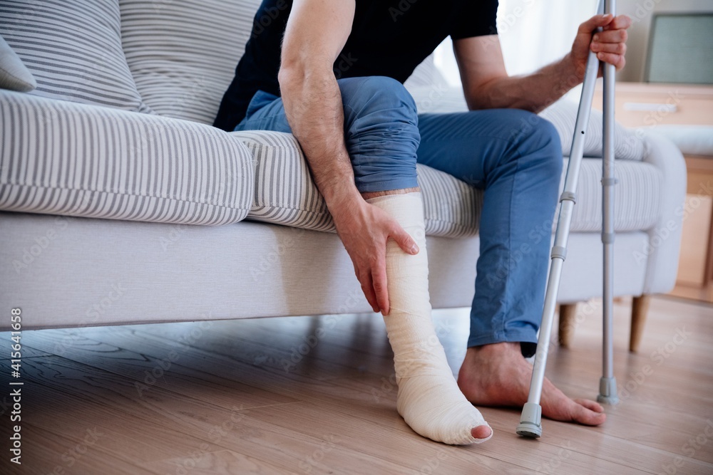 A young man with a broken leg sits on a sofa with a white plaster on his leg after an accident. He holds crutches in his hand.