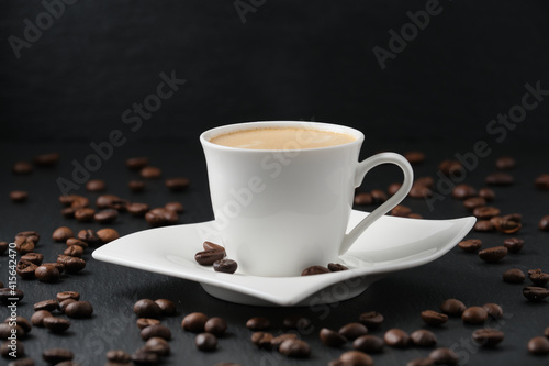 Coffee beans on a black table. White cup of coffee on a black background. Close-up side view.
