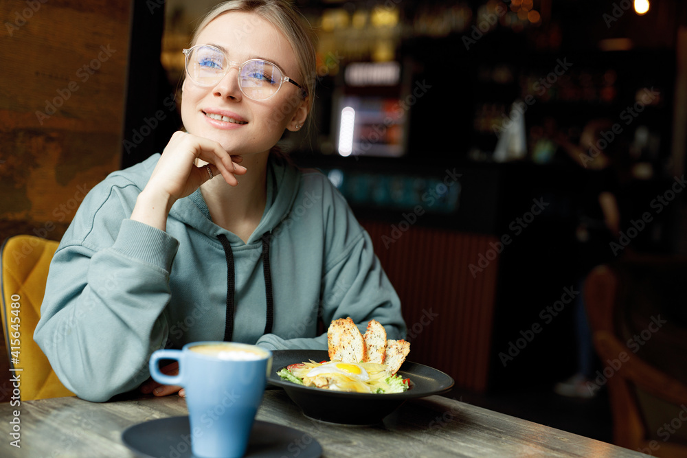 Beautiful smiling female in cafe drink coffee and eating salad. Young woman in casual clothes and glasses.