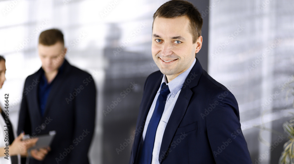 Portrait of a self-confident middle aged businessman in a blue suit, standing in a modern office with his colleagues at the background. Concept of business success Portrait of a self-confident middle aged businessman in a blue suit, standing in a modern office with his colleagues at the background. Concept of business success