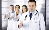 Group of professional doctors are standing as a team with thumbs up in a hospital office, ready to help their patients. Medical help, insurance in health care and medicine concept