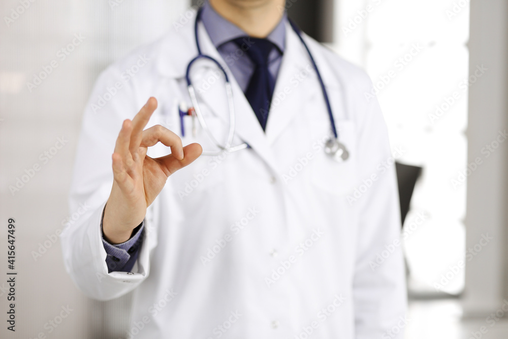 Unknown male doctor standing straight with OK sign in clinic near his working place, closeup. Perfect medical service in hospital. Medicine and healthcare concept Unknown male doctor standing straight with OK sign in clinic near his working place, closeup. Perfect medical service in hospital. Medicine and healthcare concept