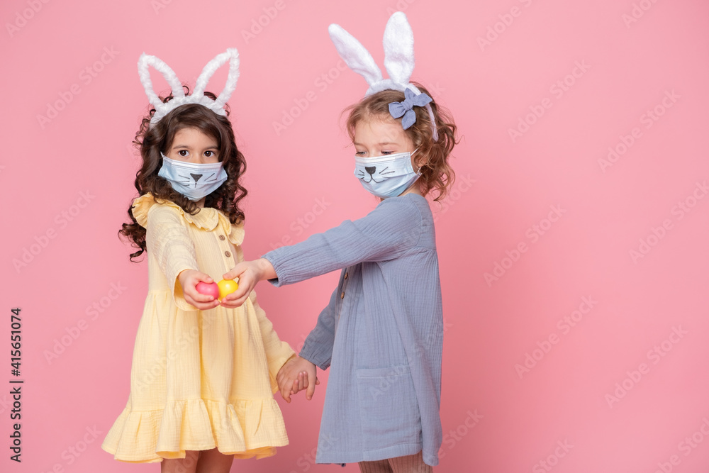two girls in bunny ears headband and protective mask with colored eggs on pink background. Covid easter concept