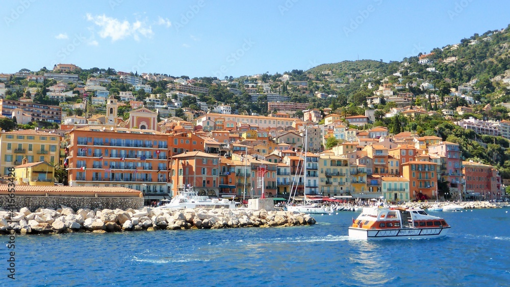 Panorama sur la ville et le port coloré de Villefranche-sur-Mer, sur la côte d’azur, dans les Alpes-Maritimes, avec un bateau naviguant sur l'eau bleue de la mer Méditerranée (France)
