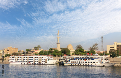 Several cruise ships lie on the banks of the Nile in the Upper Egyptian city of Luxor. In the background is the city with buildings and a minaret.