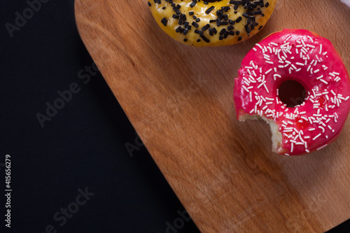 Bitten pink donut on a wooden table with a yellow donut on a black background.