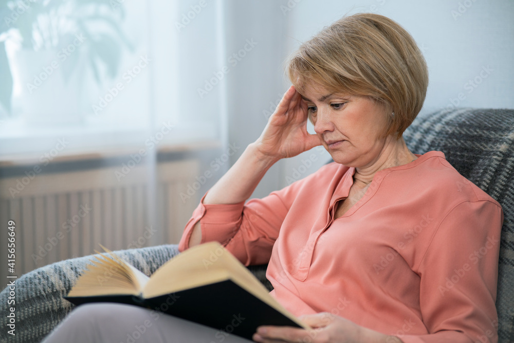 Portrait of beautiful intelligent adult senior woman is reading interesting book at home sitting on coach in living room. Portrait of beautiful intelligent adult senior woman is reading interesting book at home sitting on coach in living room.