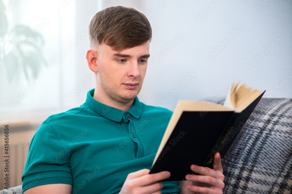 Portrait of handsome intelligent guy, young attractive serious man reading an interesting book at home sitting at couch in living room. Reader holding a book in hands. Education, leisure concept Portrait of handsome intelligent guy, young attractive serious man reading an interesting book at home sitting at couch in living room. Reader holding a book in hands. Education, leisure concept
