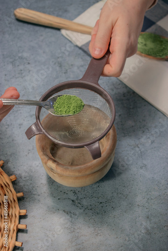 Vertical image of a matcha tea in powder on top of a small kitchen spoon with a strainer on gray stone background. Matcha tea preparation 2021.