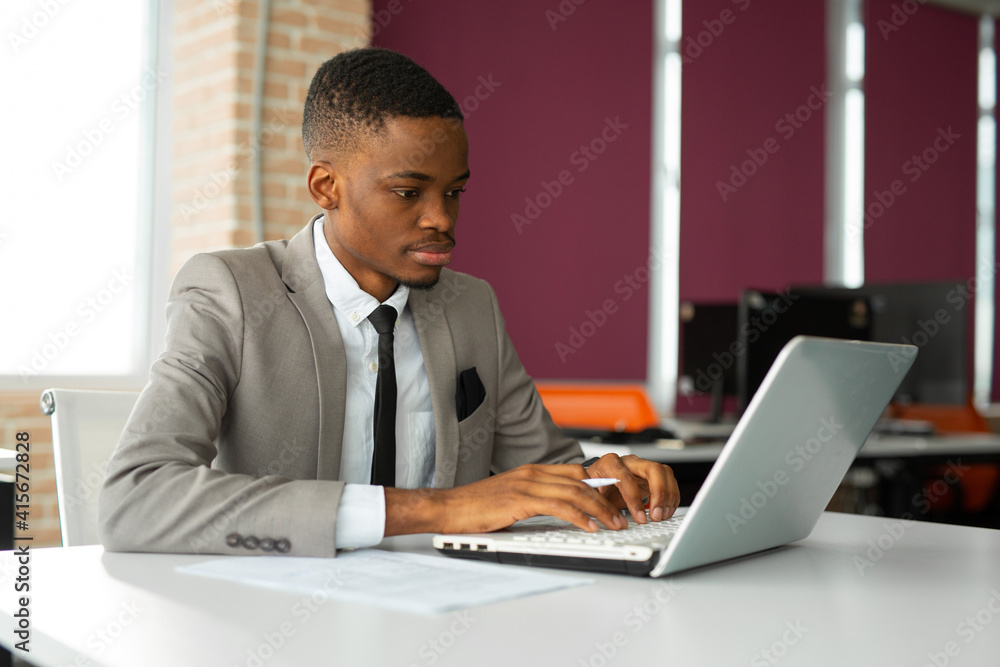 handsome young african man in suit at table in office with laptop handsome young african man in suit at table in office with laptop