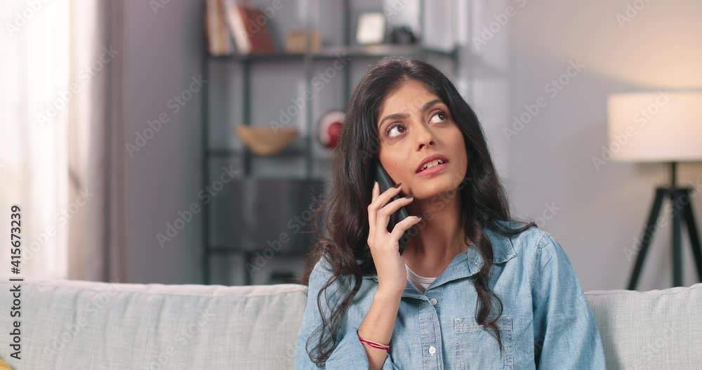 Close up of beautiful young Hindu woman sitting on sofa in living room in apartment and speaking on cellphone, pretty female calling on smartphone spending time at home, communication concept Close up of beautiful young Hindu woman sitting on sofa in living room in apartment and speaking on cellphone, pretty female calling on smartphone spending time at home, communication concept