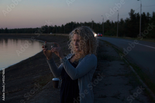Cute young girl with curly blond hair taking photo of scenic seaside during sunset