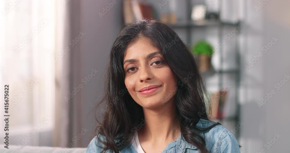 Close up of cheerful beautiful young Hindu female looking at camera and smiling in positive mood at home. Pretty dark-haired woman in good mood in living room, happy face, emotions concept Close up of cheerful beautiful young Hindu female looking at camera and smiling in positive mood at home. Pretty dark-haired woman in good mood in living room, happy face, emotions concept