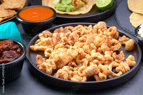Mexican pork rinds (chicharron) on a black plate, in the background Mexican food like a chicharron taco, tortillas, tortilla chips and sauces to go with it.