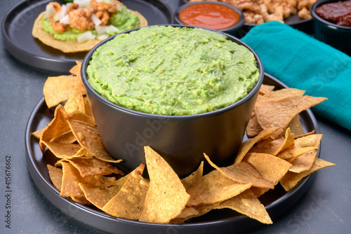 Mexican guacamole with a side of tortilla chips on a black bowl. in the background Mexican food like a pork rind taco, tortillas, pork rind on a plate and salsas to go with it.in close up photo