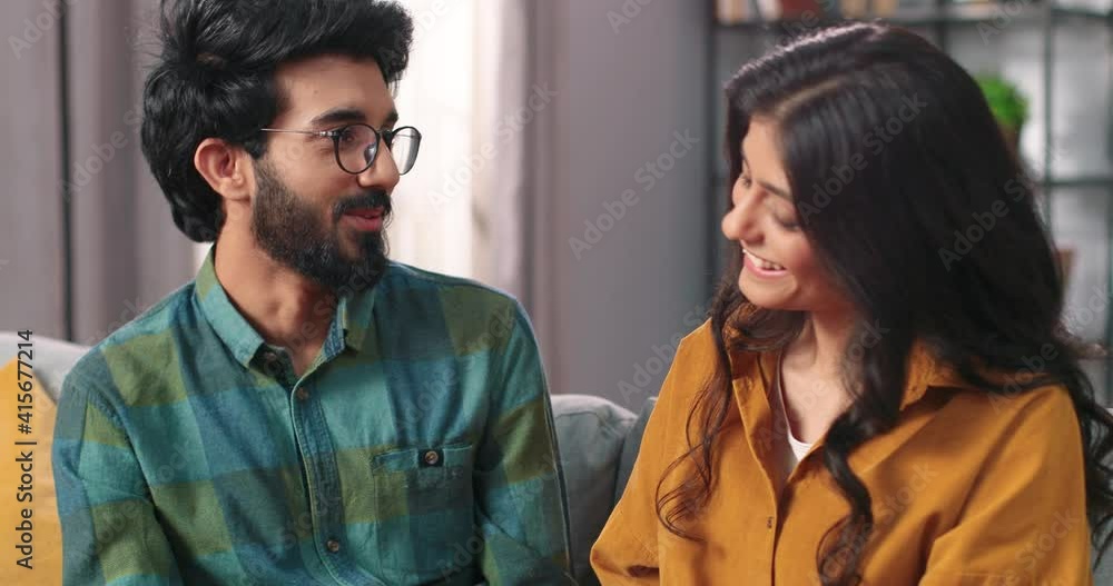 Close up of happy caring Hindu young man in good mood speaking with beautiful beloved woman sitting on sofa in cozy room at home holding hands together, cute couple, love relations concept Close up of happy caring Hindu young man in good mood speaking with beautiful beloved woman sitting on sofa in cozy room at home holding hands together, cute couple, love relations concept