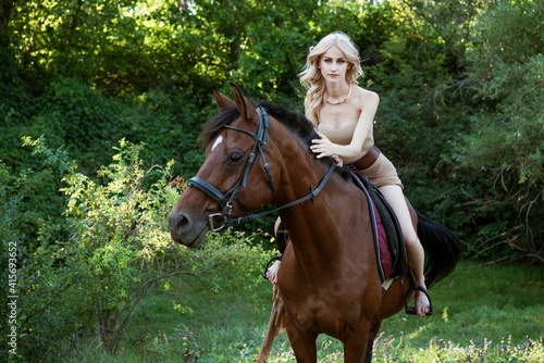 Beautiful blonde woman riding a horse in a dress outdoors in a summer park