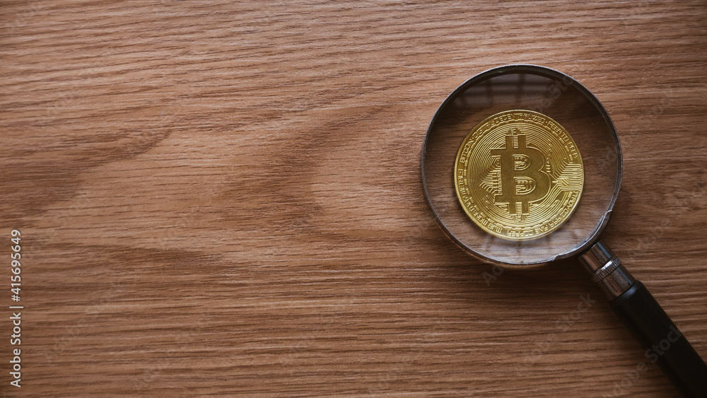 A magnifying glass magnify the replica of gold bitcoin on wooden background with copy space. A magnifying glass magnify the replica of gold bitcoin on wooden background with copy space.