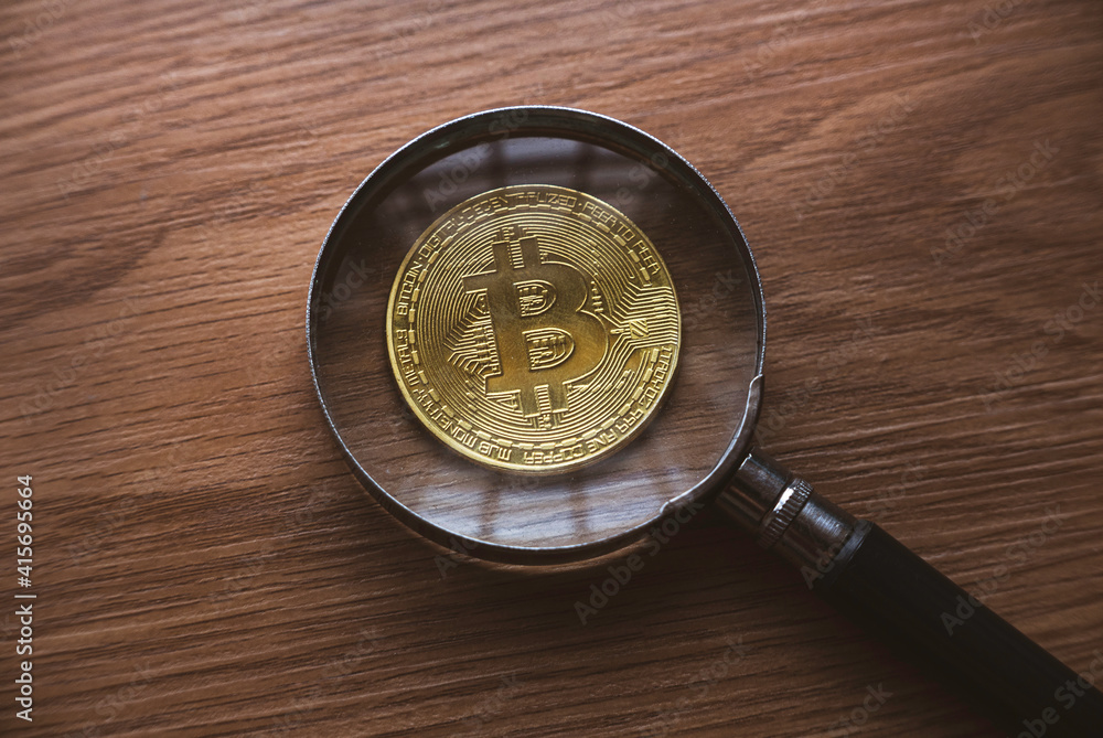 A magnifying glass magnify the replica of gold bitcoin on wooden background.