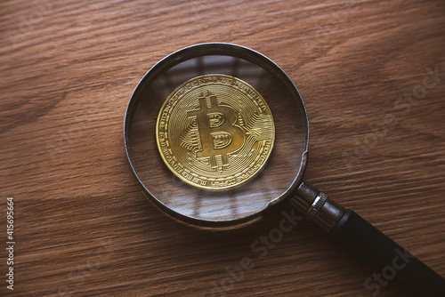 A magnifying glass magnify the replica of gold bitcoin on wooden background.