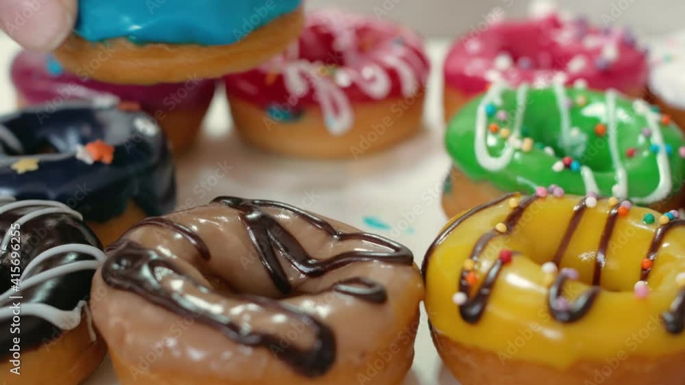 With two fingers of the brush, take one donut with blue glaze. Donuts are doused with fruit glaze of different colors and dark white chocolate. Sweets close-up. Junk food. With two fingers of the brush, take one donut with blue glaze. Donuts are doused with fruit glaze of different colors and dark white chocolate. Sweets close-up. Junk food.