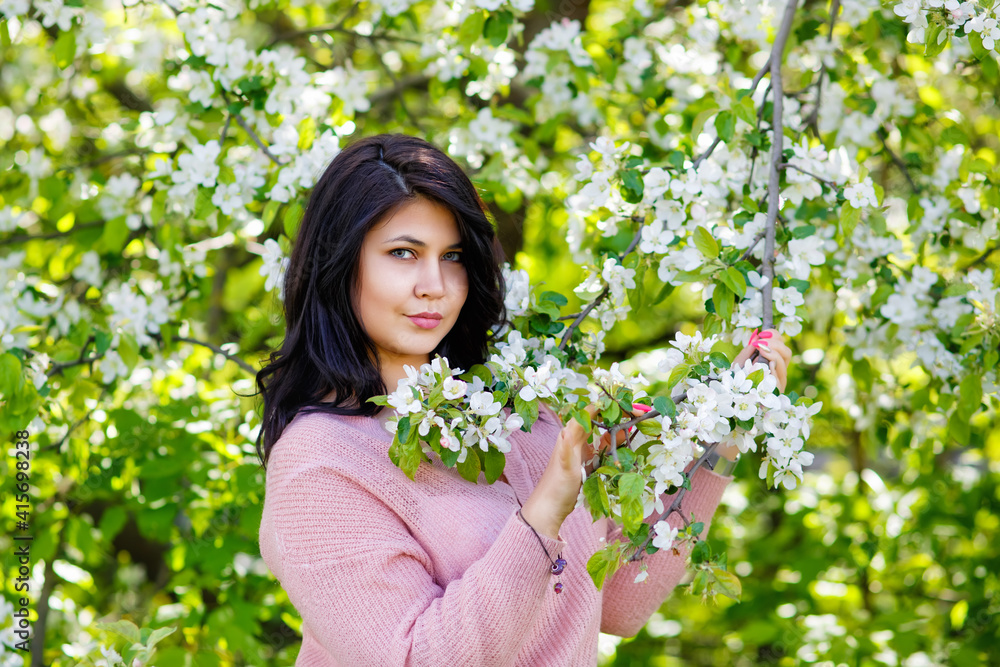 Portrait of a fat plump young woman with black hair and a blooming apple tree on a white floral background in a park on a spring day. Happy girl plus size. Portrait of a fat plump young woman with black hair and a blooming apple tree on a white floral background in a park on a spring day. Happy girl plus size.