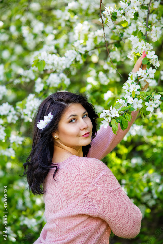 Portrait of a fat plump young woman with black hair and a blooming apple tree on a white floral background in a park on a spring day. Happy girl plus size. Portrait of a fat plump young woman with black hair and a blooming apple tree on a white floral background in a park on a spring day. Happy girl plus size.