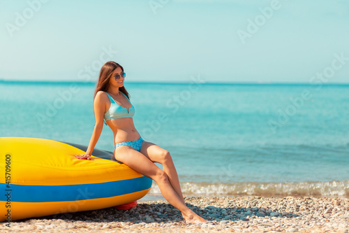 A beautiful woman in sunglasses and a bikini poses leaning on an inflatable boat. In the background, the sea and sky. Concept of beach holidays and summer vacations