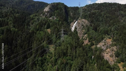 Power Cables Over The Italian Alps Surrounded By Green Woodland With Waterfall On The Background. aerial