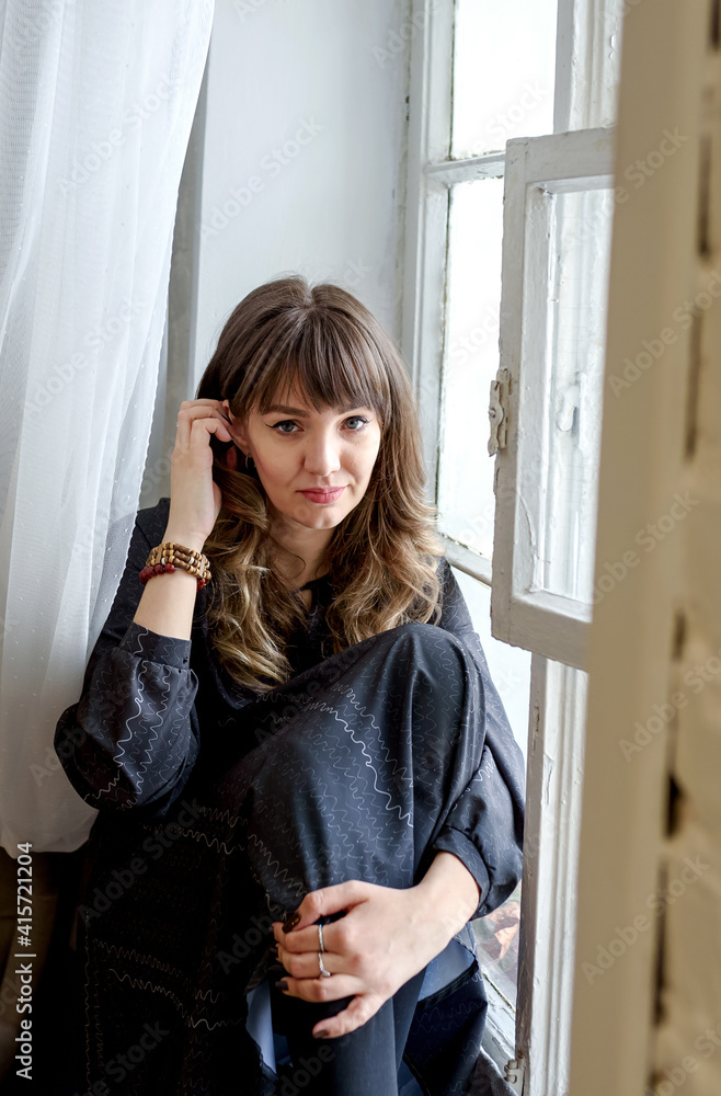 Young brunette woman in a dress sits dreamily by the window