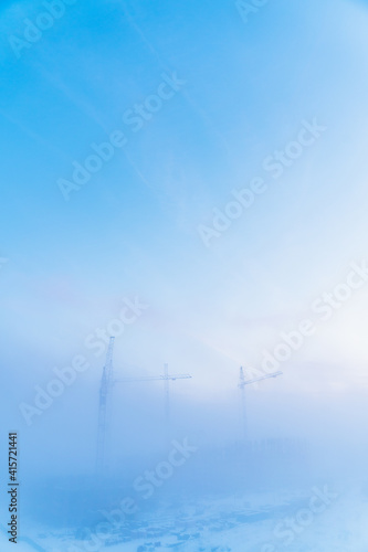Tall tower cranes shine through in the winter morning fog. Bird's-eye view of the construction site on a foggy morning.