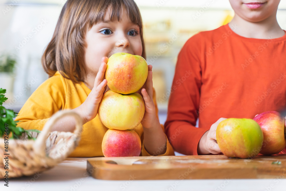 Fototapeta premium Funny kids building pyramid of apples in kitchen, crazy having fun. Happy sister and brother cook together laughing, healthy juices and fresh fruit salad. Fun vegan family cooking events