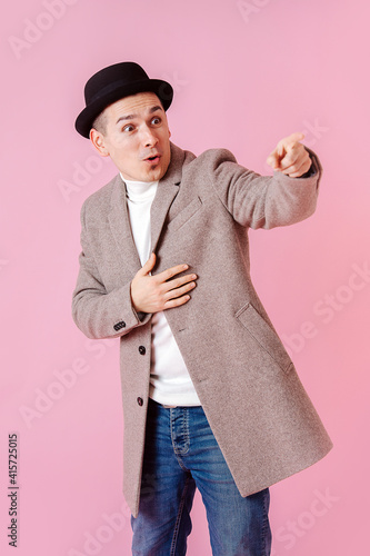 Alert emotional man dressed in a grey beige coat and a classical porkpie hat points at something. Over dark pink background. photo