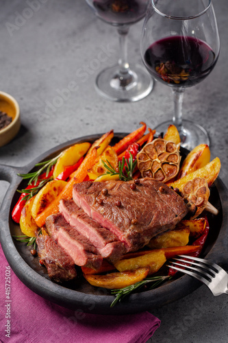 Steak with baked vegetables and red wine glases. Vertical image, grey background, copy space.